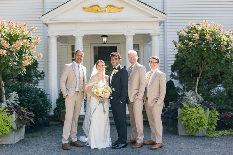 Bride and groom posing with wedding party in light tan suits outside a white-columned venue entrance.