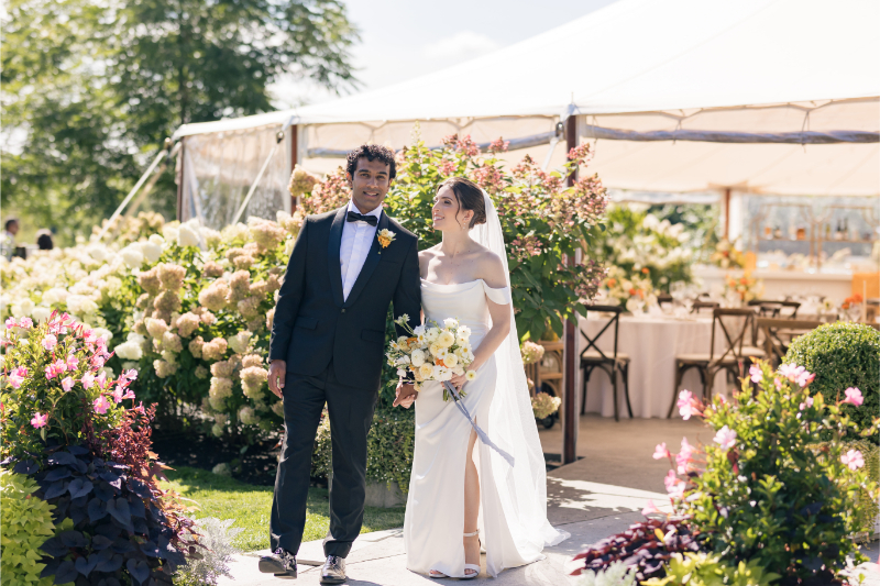 Bride and groom walk hand in hand through lush garden blooms outside their tented wedding reception.