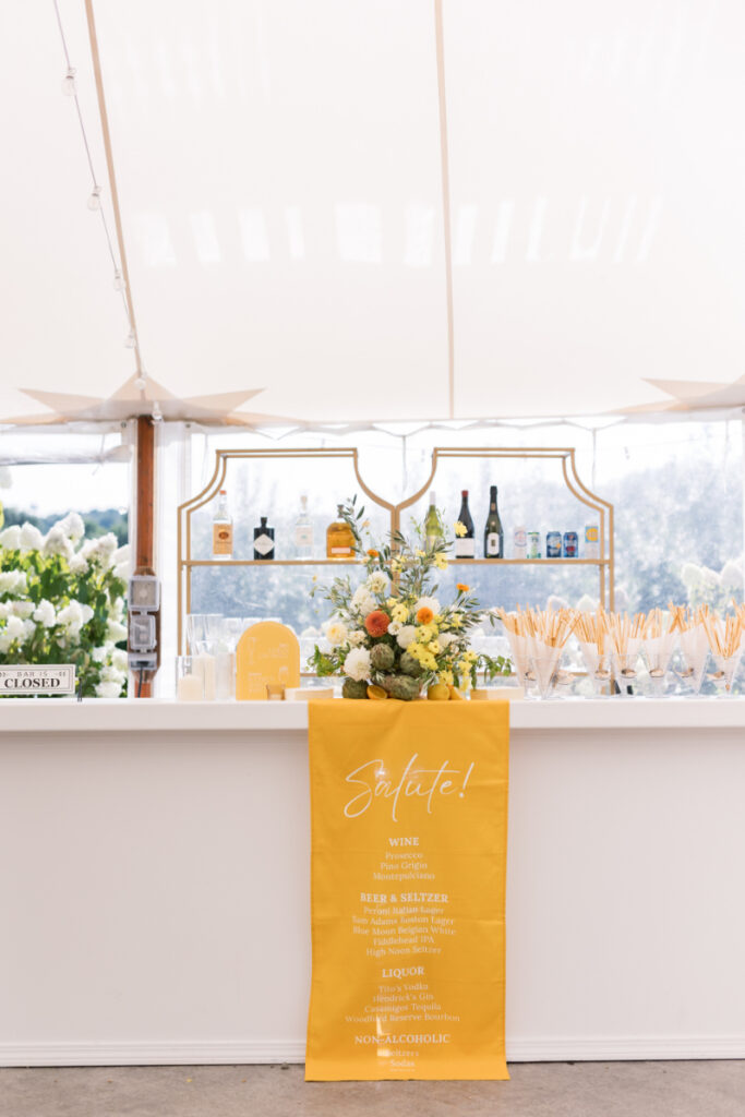 White bar setup with yellow cocktail menu, citrus florals, and grissini under a sailcloth tent.