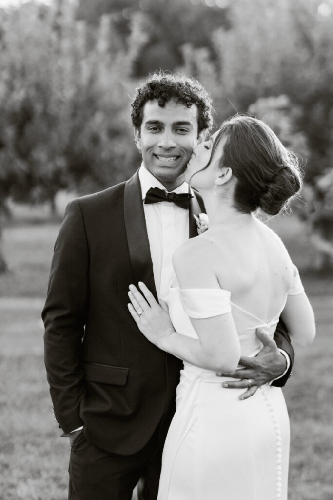 Black and white photo of bride kissing groom’s cheek while he smiles during a joyful wedding moment.