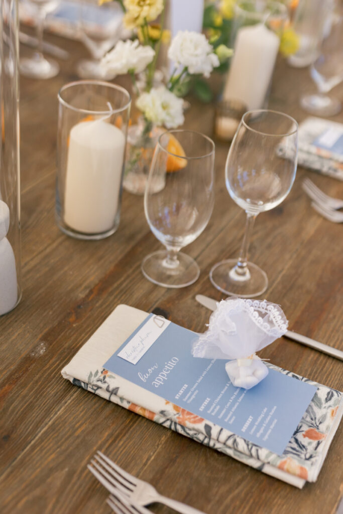 Close-up of place setting with floral napkin, blue menu card, and favor on a wooden table.