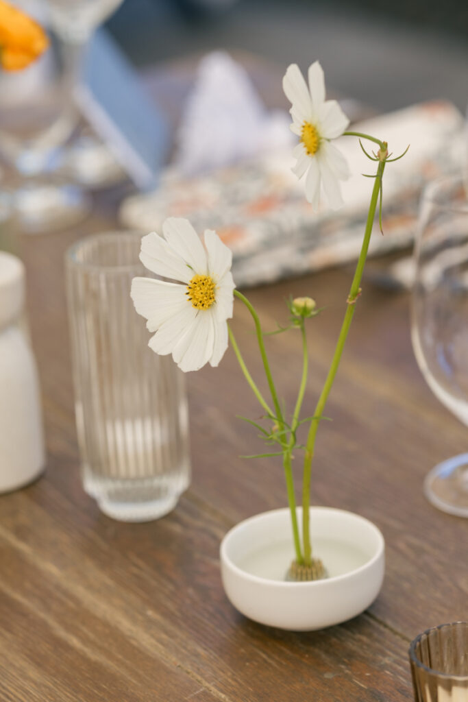 Minimalist bud vase with white cosmos flower on a wooden reception table with textured glassware.