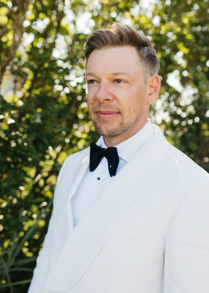 Groom portrait in white tuxedo during elegant outdoor Block Island wedding surrounded by lush coastal greenery.