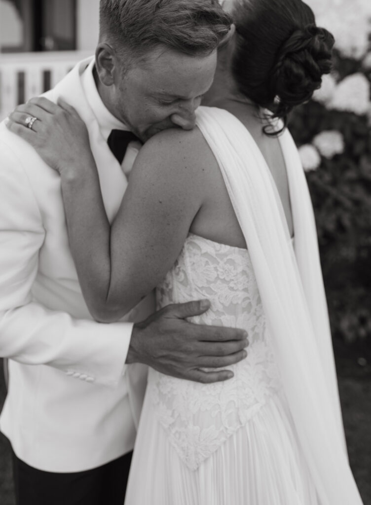 Groom embracing bride in lace gown during intimate black and white Block Island wedding portrait.