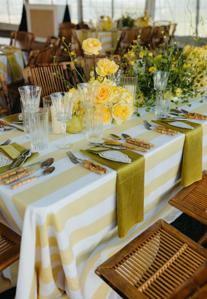 Reception table with yellow striped linens, roses, pears, and bamboo flatware inside tent.