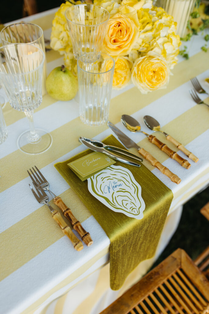 Place setting with oyster shaped menu, striped linens, and yellow floral centerpiece.