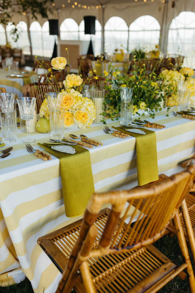 Reception table with yellow striped linens, roses, greenery, and bamboo chairs inside tent.