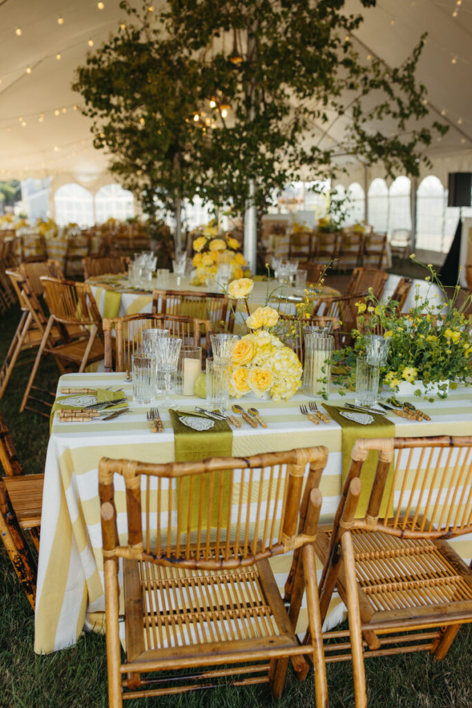 Reception tables with yellow striped linens, bamboo chairs, and lush greenery under tent canopy.
