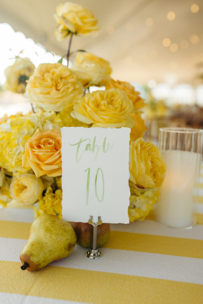 Yellow rose centerpiece with handwritten table number card and pear detail on striped linen table.