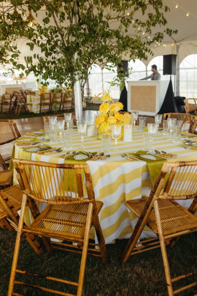 Round reception table with yellow striped linens and bamboo chairs under greenery filled tent.