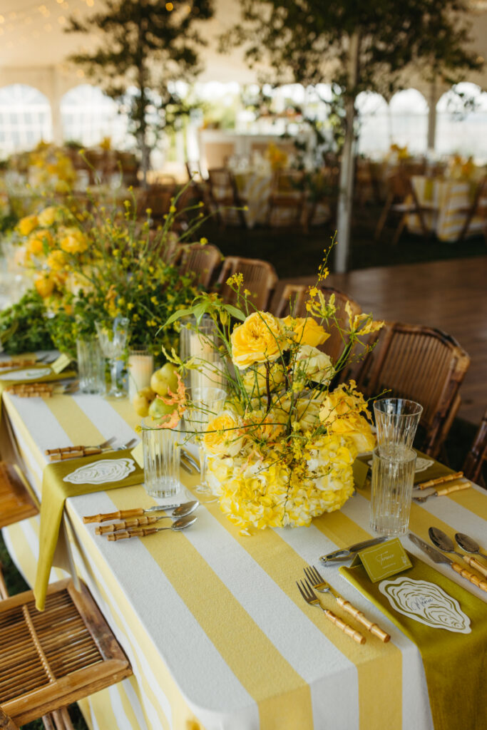 Round reception table with yellow striped linens and bamboo chairs under greenery filled tent.