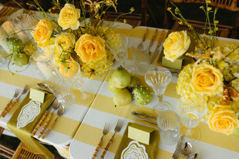 Reception table with yellow striped linens, roses, pears, crystal glassware, and bamboo flatware details.