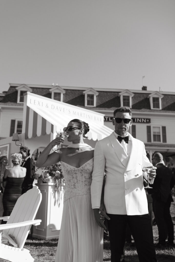 Bride sipping cocktail beside groom near striped bar during outdoor wedding celebration.
