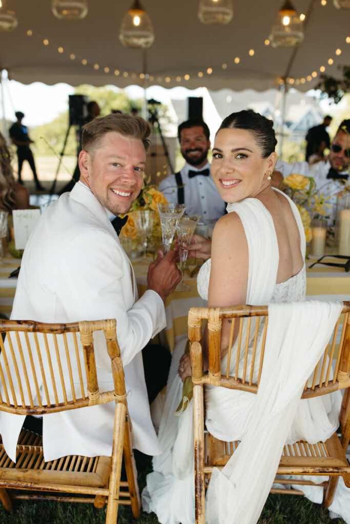 Bride and groom smiling at reception table under tent lights, seated in bamboo chairs.