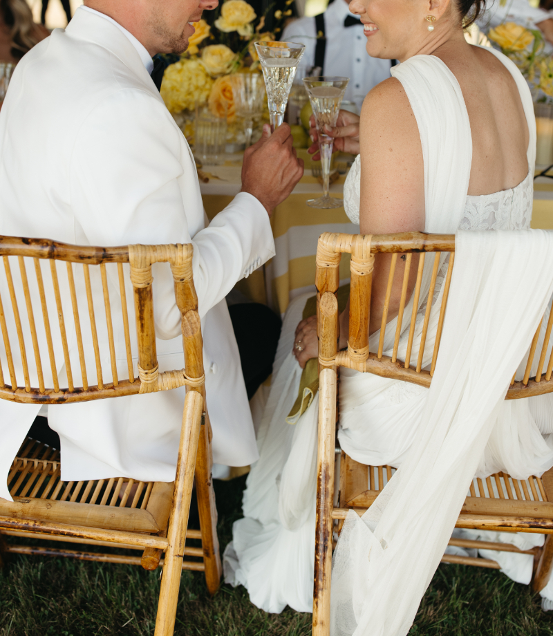 Newlyweds toast with champagne at reception table, bride’s draped veil flowing over bamboo chair.