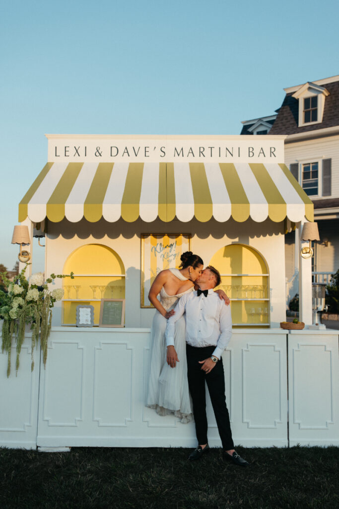 Bride kissing groom while standing behind yellow striped martini bar during sunset cocktail hour moment.