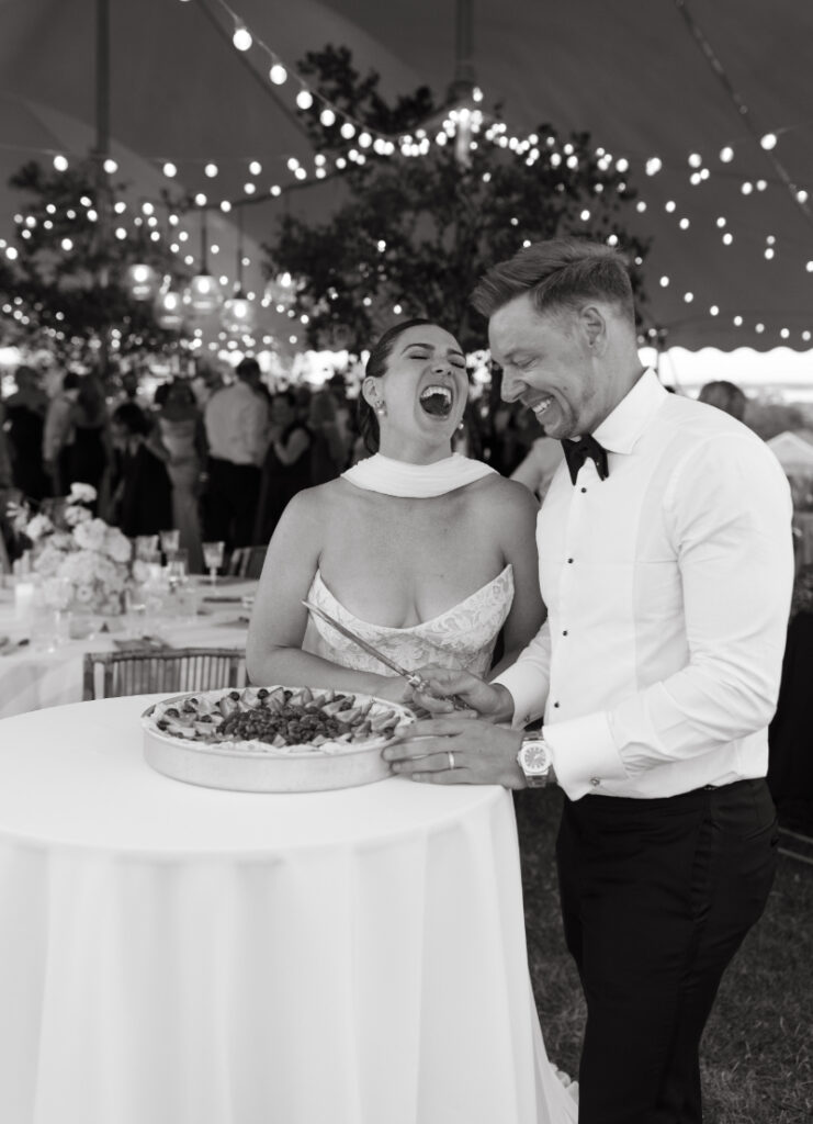 Bride and groom laughing while cutting cake under glowing string lights during evening reception.