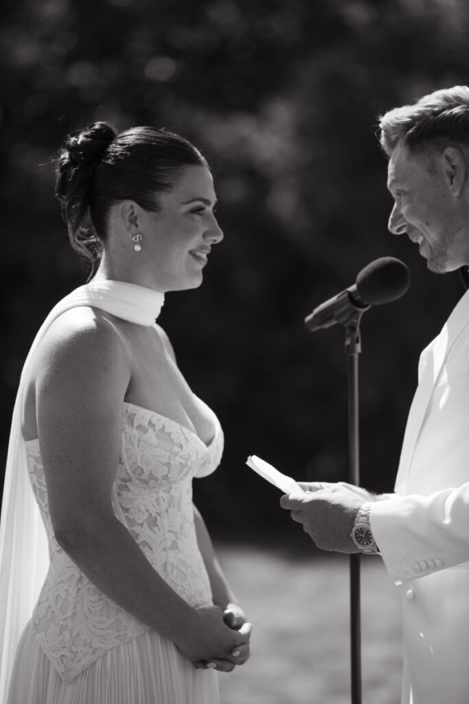 Bride and groom smiling while reading vows to each other during outdoor wedding ceremony.