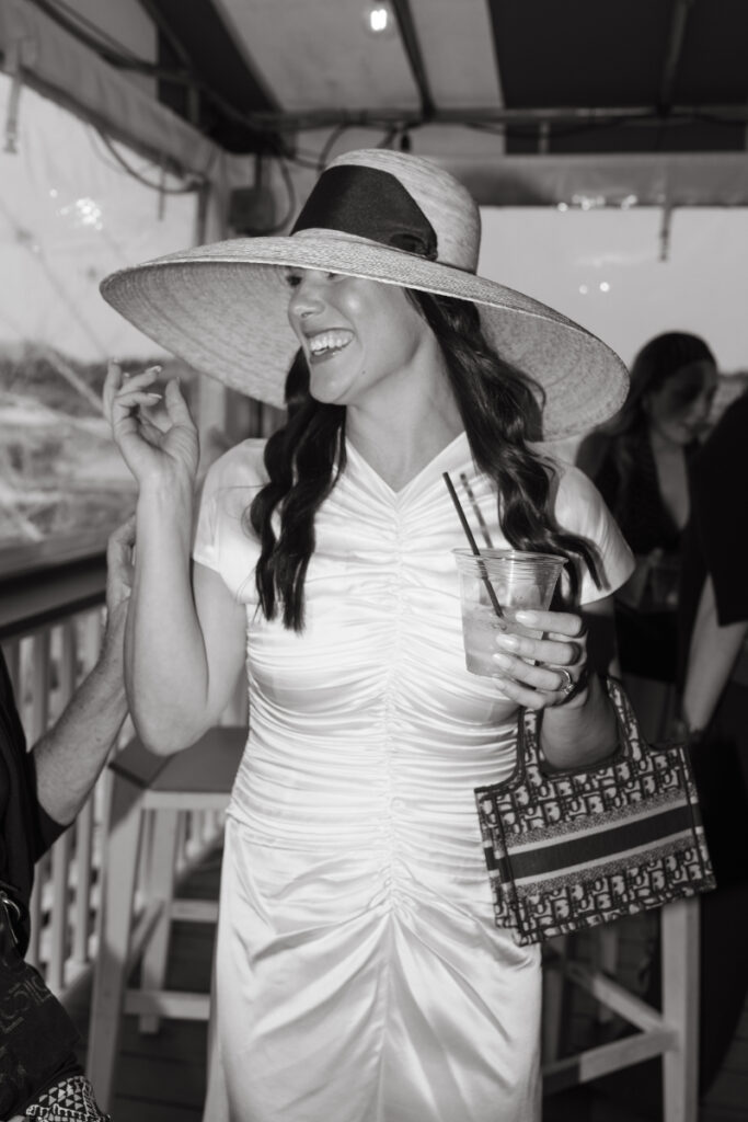 Bride laughing in wide brim hat while holding cocktail during stylish Block Island wedding welcome party.