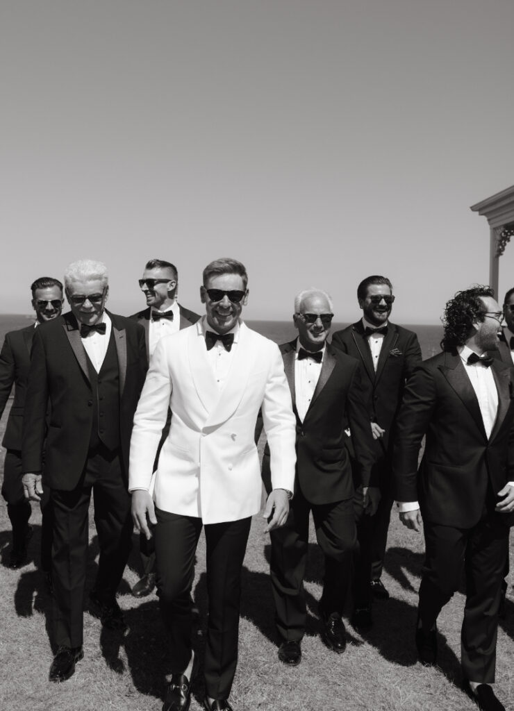 Groom and groomsmen posing by the ocean in black tuxedos and white dinner jacket.