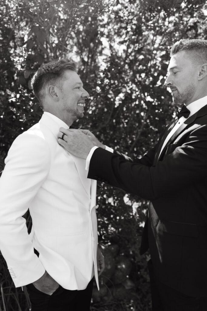 Groomsman adjusting groom’s bow tie and white dinner jacket during pre-ceremony preparations.
