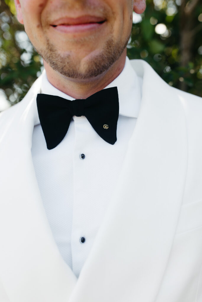 Close-up of groom wearing classic black bow tie with white tuxedo jacket.