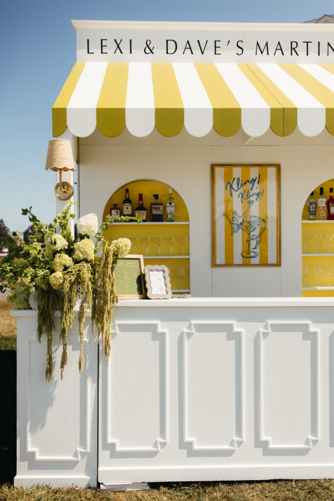 Custom martini bar with yellow striped awning and floral details during outdoor wedding cocktail hour.