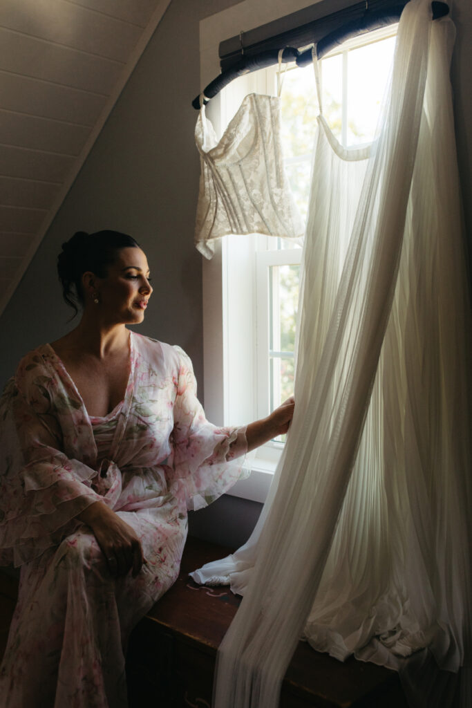 Bride sitting by window holding her wedding dress and veil during a Block Island Wedding