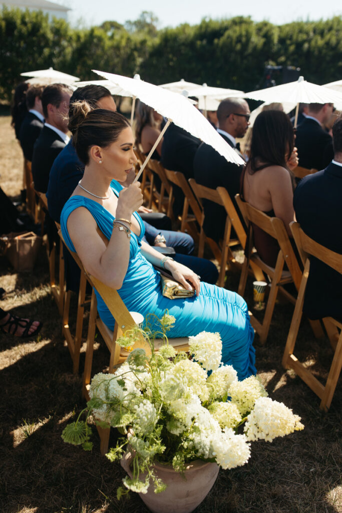 Guest seated under parasol during sunny outdoor ceremony with hydrangea floral arrangements.