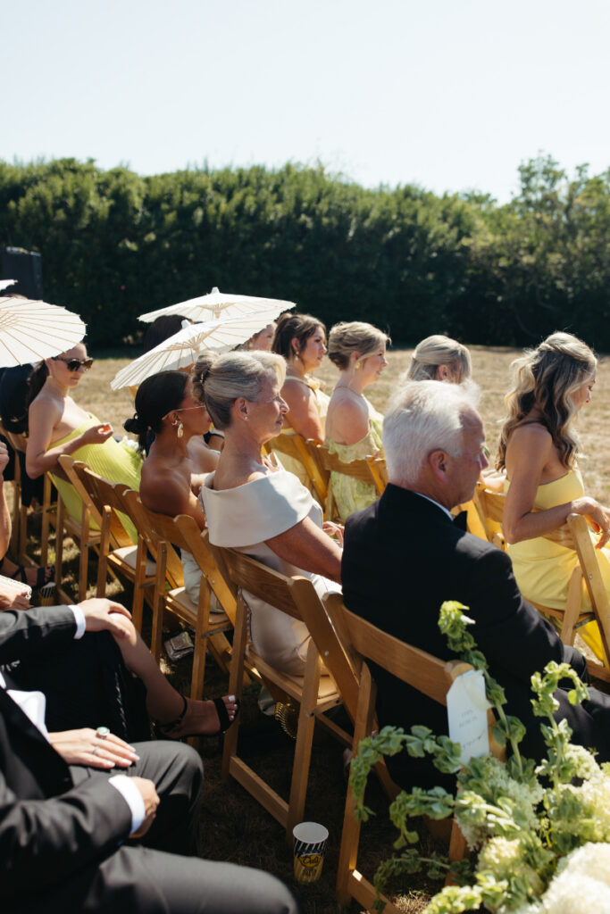 Wedding guests seated in wooden chairs holding parasols during warm outdoor ceremony.