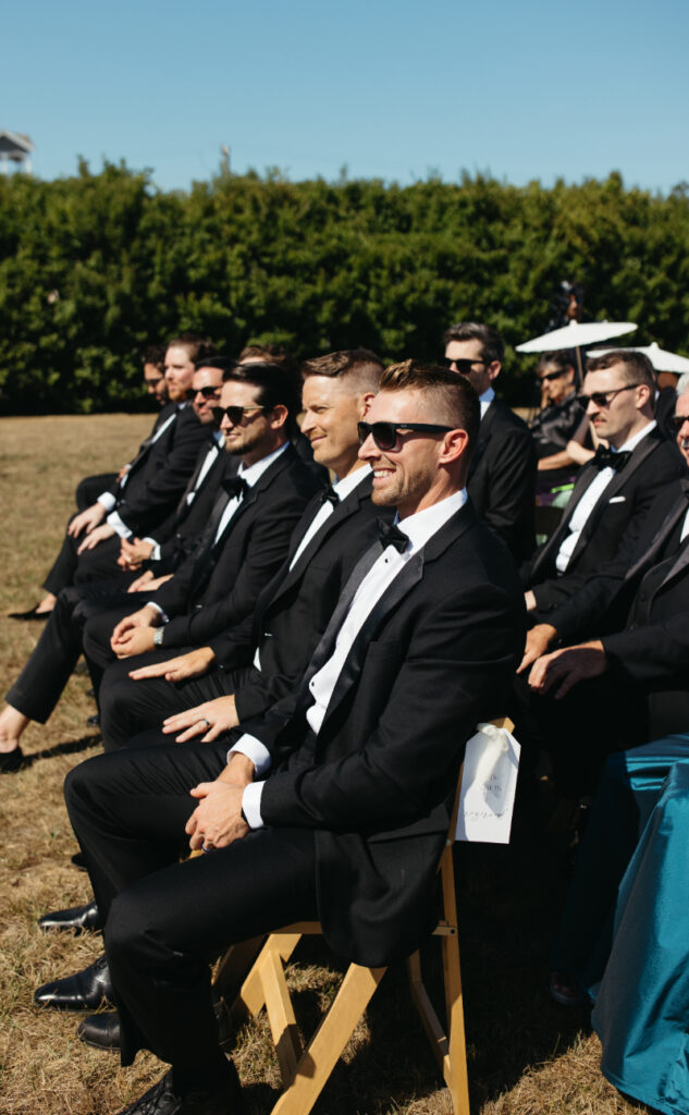 Groomsmen seated together in black tuxedos smiling during outdoor ceremony.
