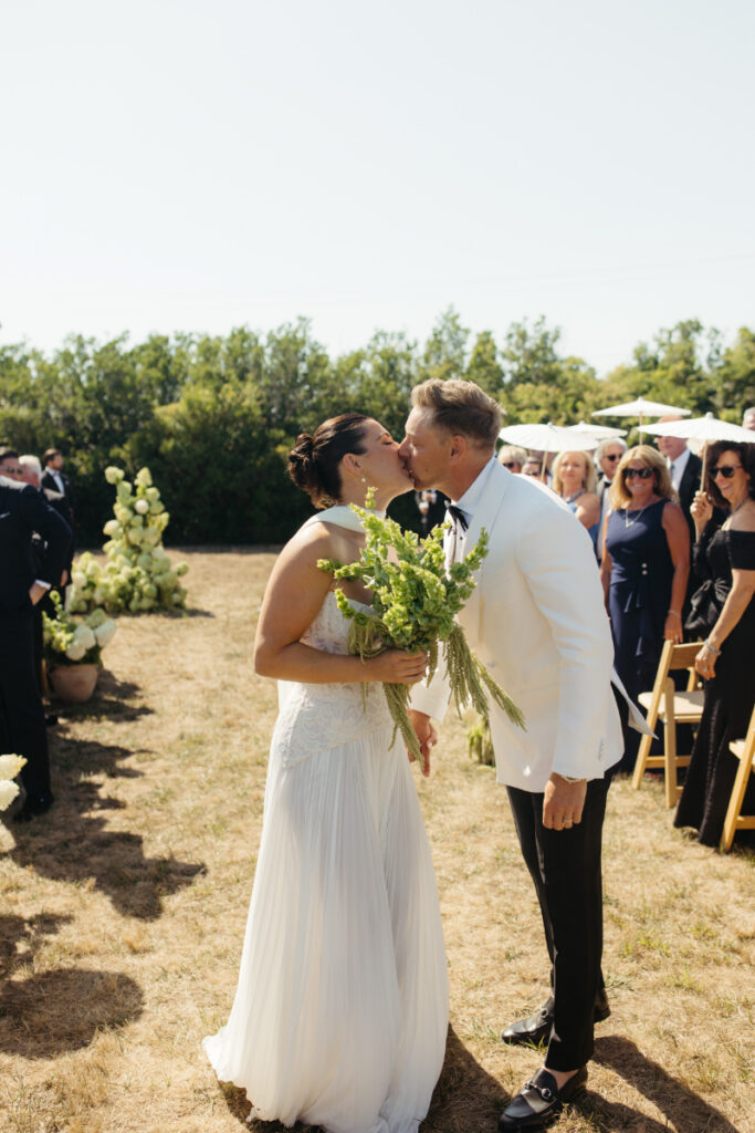 Bride and groom kissing after ceremony while guests stand and celebrate behind them.