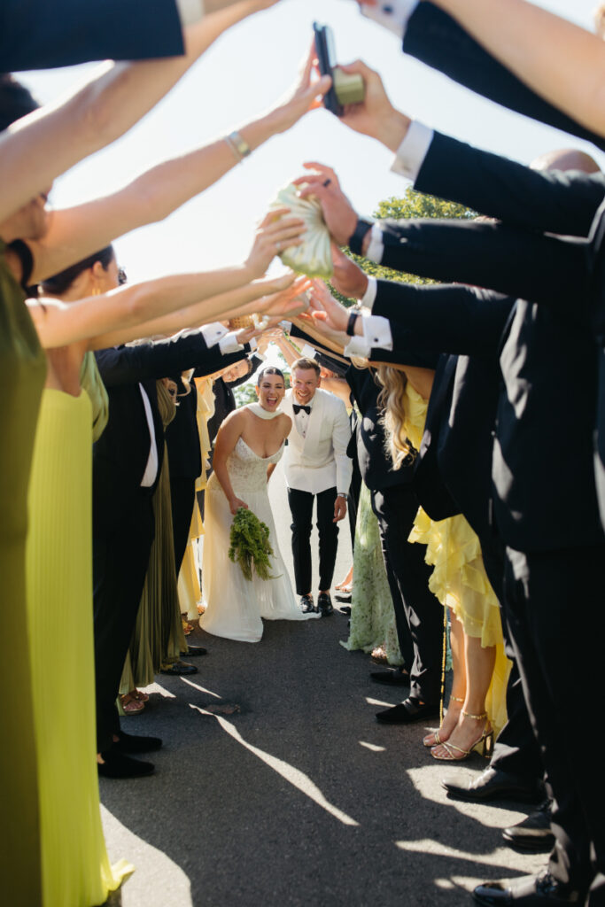 Newlyweds walking through celebratory champagne spray as guests cheer and raise bottles.