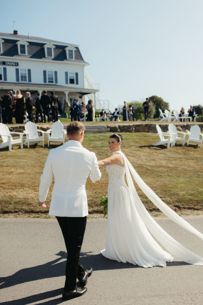 Bride leading groom across lawn toward reception as guests gather nearby.