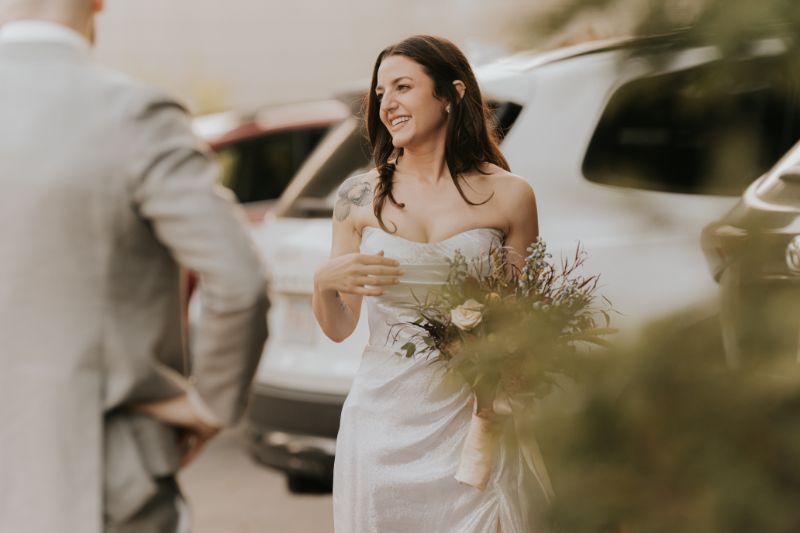 Bride smiling in strapless satin gown holding colorful bouquet outdoors.