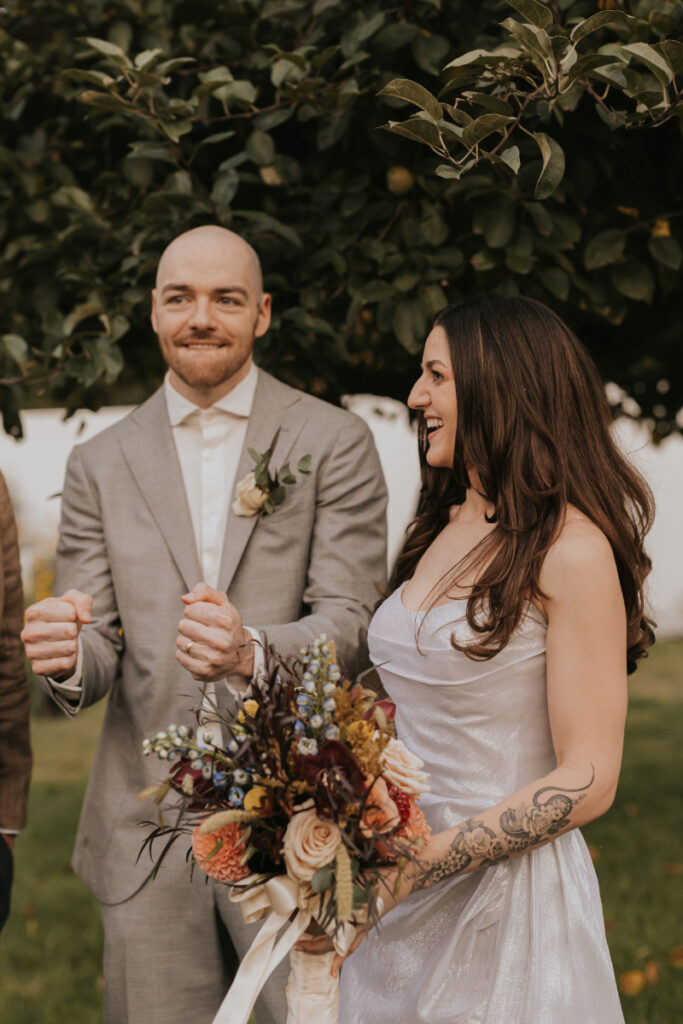 Couple smiling outdoors; bride with bouquet, partner in light gray suit.