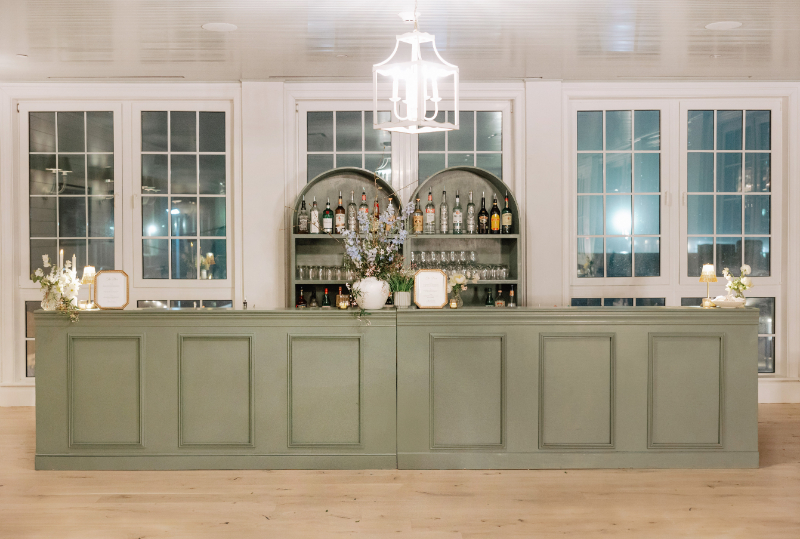 Green built-in bar with arched shelving, stocked with glassware and bottles, styled with a floral arrangement and framed menu