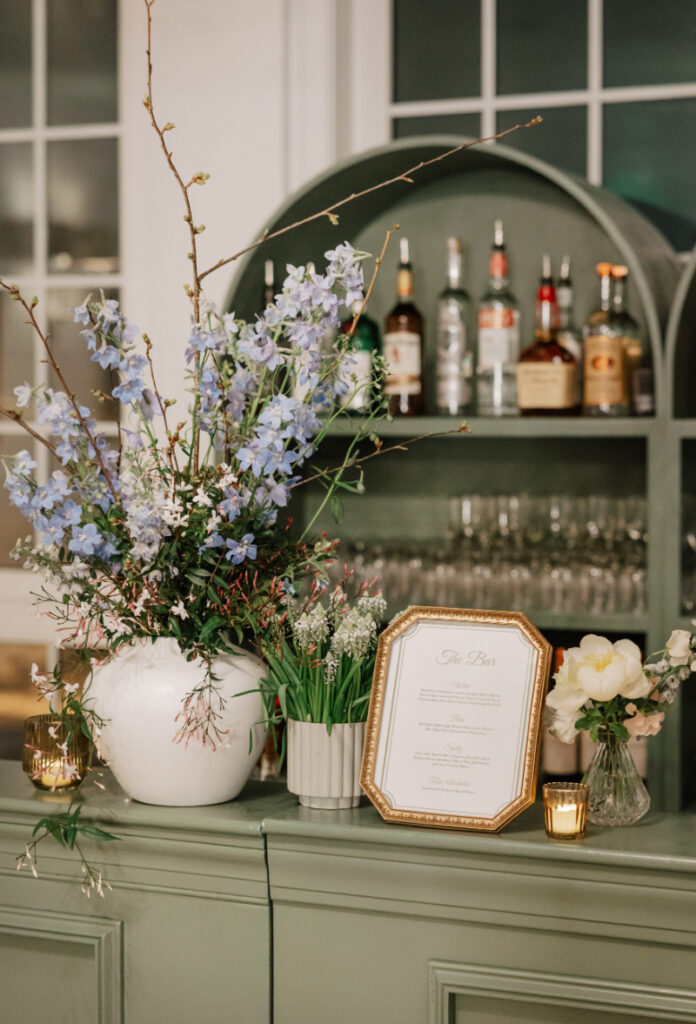 Close-up of bar styling with blue and white florals, glassware, and a framed drink menu on a green bar counter