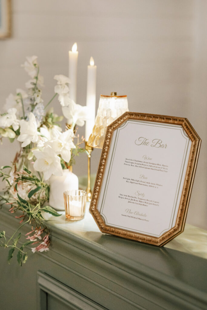 Framed cocktail menu on a green bar with candles and white floral arrangements nearby