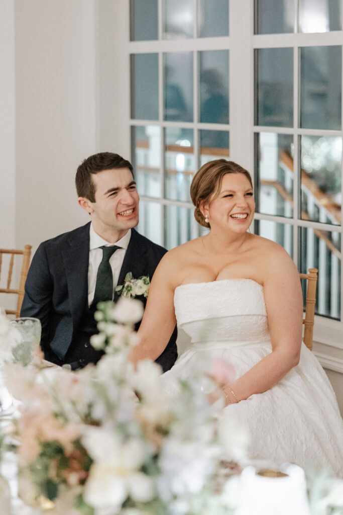 Couple seated side by side at their reception table smiling and looking toward a speaker