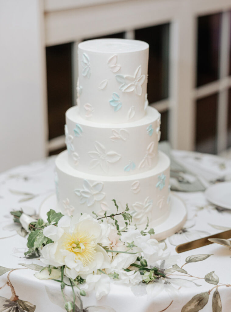 Three-tier white wedding cake with floral detailing and fresh flowers at the base on a patterned linen table