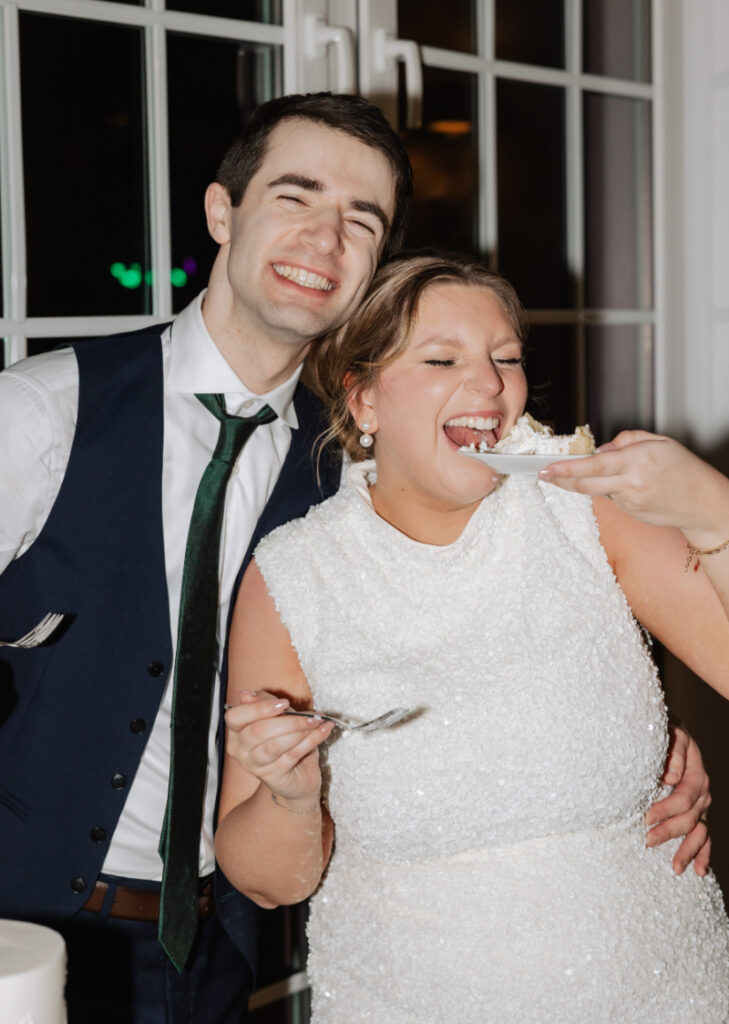 Bride laughing while being fed a bite of cake by her partner during the cake cutting celebration