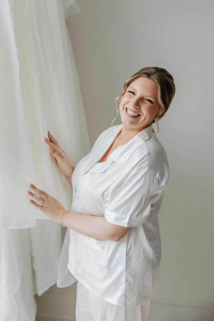 Bride smiling in getting ready outfit beside wedding dress, soft morning moment by Newport wedding planner
