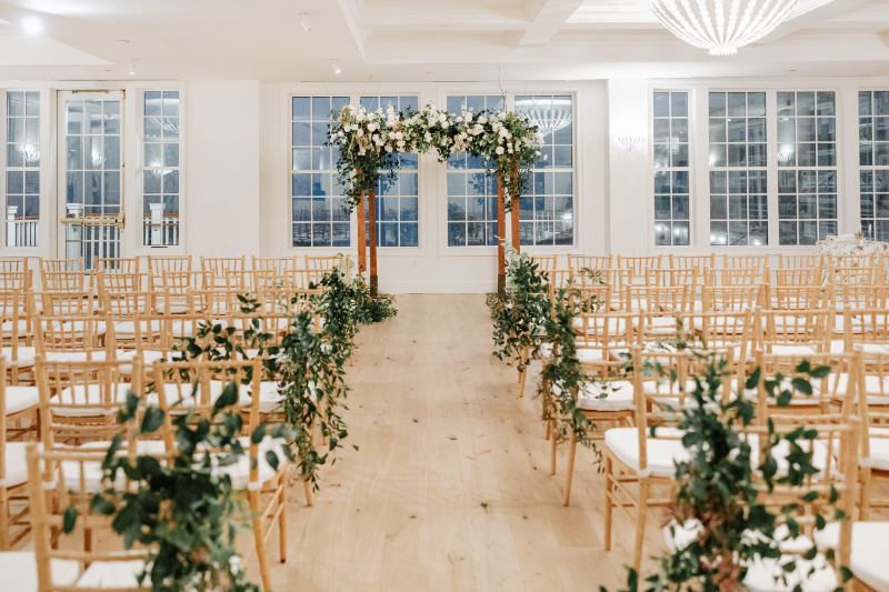 Indoor ceremony aisle with greenery-lined chairs and floral arch, romantic Newport wedding ceremony setup