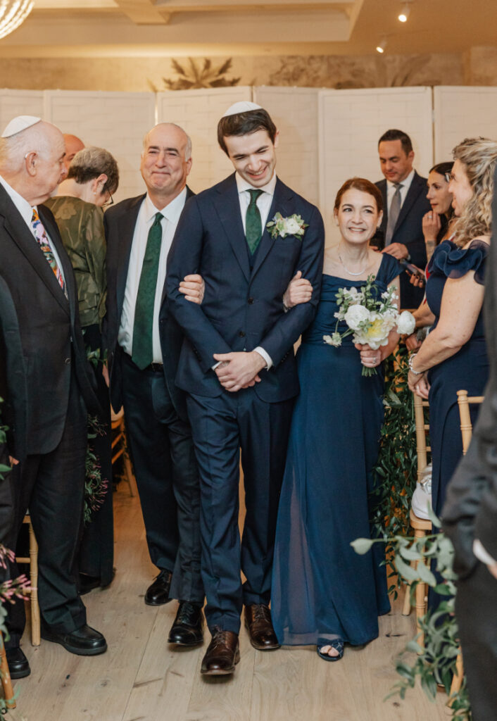 Groom walking down aisle with family, smiling candid moment before ceremony begins