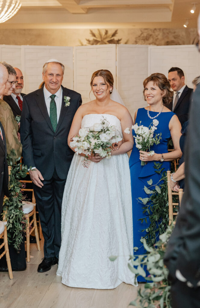 Bride entering ceremony with parents, emotional aisle moment surrounded by guests and greenery