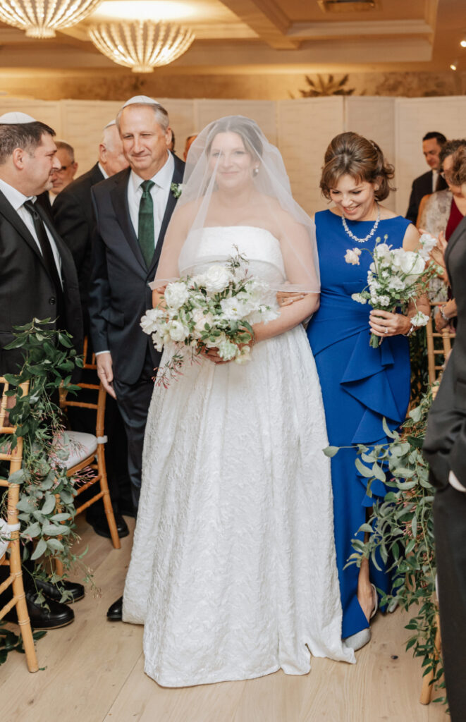 Bride walking down aisle with parents, soft smile and bouquet as guests look on