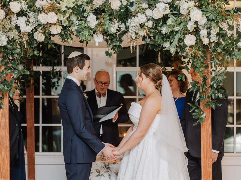 Couple holding hands under floral chuppah, exchanging vows during heartfelt ceremony moment