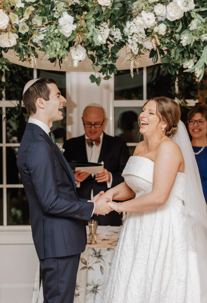 Couple laughing together during vows, joyful candid moment under lush greenery-covered ceremony structure