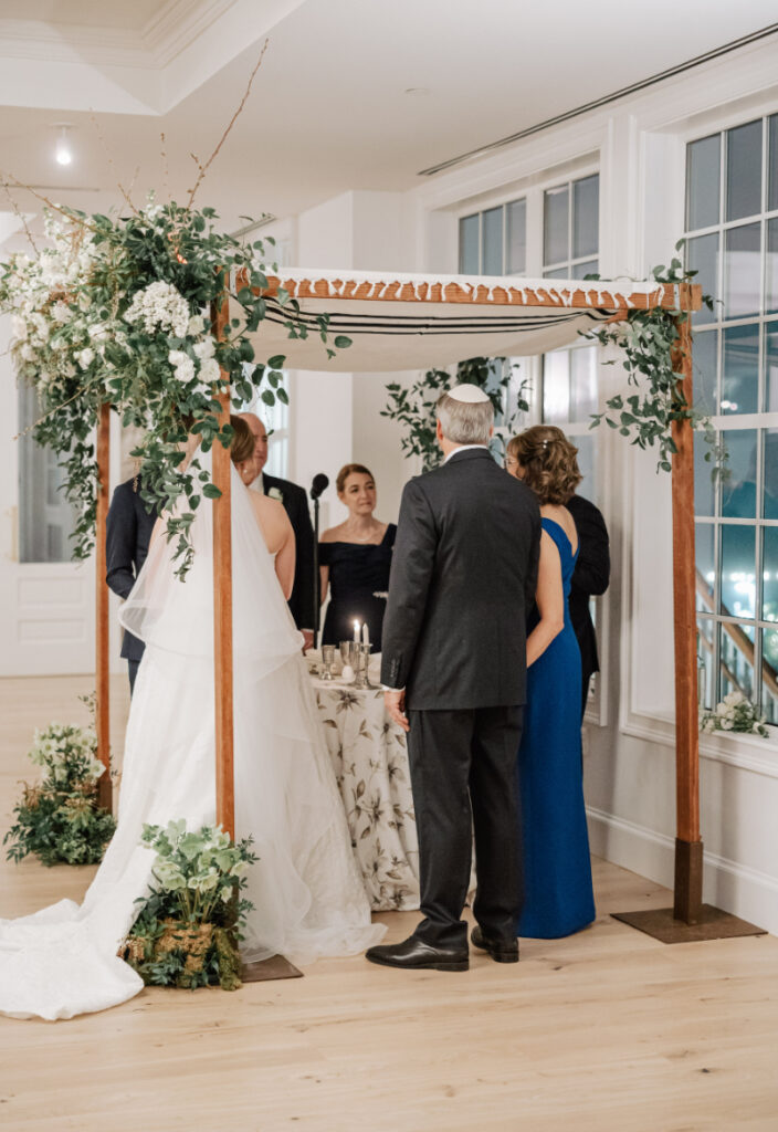 Wide ceremony view under floral canopy with family gathered, meaningful tradition captured from behind